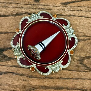 May include: A silver-toned bottle stopper with a decorative top featuring a deep red center and clear crystals, resting on a circular base with a deep red enamel finish, gold-toned metal accents, and embedded amber and clear crystals.