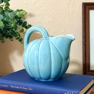 May include: A light blue ceramic pitcher shaped like a pumpkin with vertical ridges and a curved handle. The pitcher features a smooth, glossy finish and a wide pouring spout. It is positioned on top of a dark blue book with visible white text.