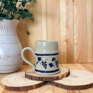 May include: A beige ceramic mug with a handle, decorated with blue grape and leaf designs. The mug has two blue bands and sits on a wooden coaster. A white vase with greenery is in the background.