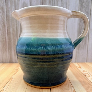 May include: A handmade ceramic pitcher with a wide spout and a curved handle. The pitcher features a gradient of colors, starting with a beige top, transitioning to teal and blue, and ending with a dark blue base. The pitcher is sitting on a wooden surface.