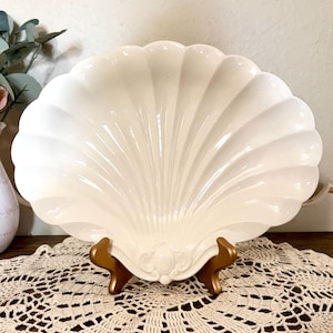 May include: A large, off-white seashell-shaped serving dish. The dish has a scalloped edge and a detailed, fan-like pattern. It is displayed on a gold-colored stand, resting on a cream-colored lace doily.