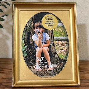 May include: A rectangular gold-toned picture frame with an oval-shaped inner opening. The frame contains a display photo of a child sitting by a tree, with text reading 5x7 Professional Portrait Frame and Deep Shadowbox Perfect for Photos Mementos Crafts.