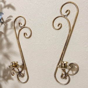 May include: A pair of gold-toned metal wall sconces featuring elongated, curved wire frames with decorative scrollwork at the top and a circular candle cup at the base of each piece. The metal has a smooth, reflective finish throughout the design.