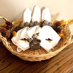 May include: A woven wicker basket containing three white cloth napkins secured with brown acorn-shaped napkin rings. The basket is decorated with artificial pinecones and yellow-tipped foliage, and one additional acorn napkin ring sits in the center.