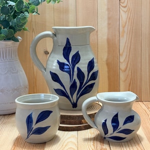 May include: A set of three ceramic pitchers and a mug. The pitchers and mug are off-white with a blue leaf design. The largest pitcher has a handle and a spout. The mug and the smallest pitcher also have handles.