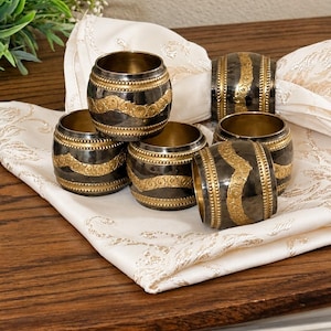 May include: A set of five cylindrical napkin rings featuring a dark metallic finish with a central band of embossed gold-toned floral patterns and beaded edges, resting on a white fabric napkin with a subtle gold-toned pattern.