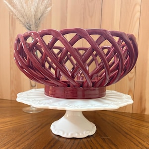 May include: A burgundy ceramic basket with a woven design sits atop a white, scalloped-edge cake stand. The basket's intricate pattern and rich color create a striking contrast against the stand's simplicity. A decorative piece for the home.