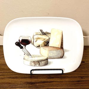 May include: White rectangular serving platter with a watercolor illustration of cheese, a glass of red wine, and a cheese knife. The platter features various cheeses, including a wrapped block and a wheel of cheese.