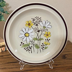 May include: A round ceramic plate with a cream-colored base and a dark brown rim. The center features a floral illustration of white daisies, yellow flowers, and green leaves with thin brown stems and small dark brown berries on slender branches.