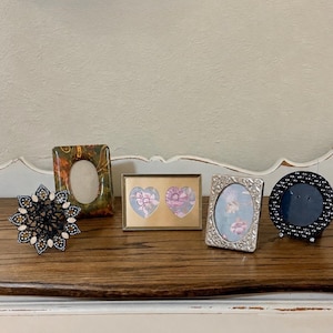 May include: A collection of four picture frames: a black star-shaped frame with white accents, an oval frame with a green and orange patterned finish, a rectangular gold frame with two heart-shaped openings, and a silver oval frame with a textured border.