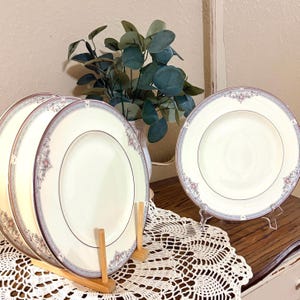 May include: A set of three round ceramic dinner plates featuring a cream-colored center and a decorative border with intricate blue, grey, and pink floral patterns. The plates are displayed upright on wooden stands, showcasing their detailed rims.