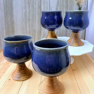 May include: Four handmade ceramic goblets. The goblets have a deep blue glaze on the bowl and a textured, tan-colored base. The goblets are arranged on a light-colored wooden surface, with one goblet on a white, decorative plate.