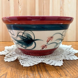 May include: A large, decorative ceramic bowl with a wide rim. The bowl features a burgundy rim, a teal band, and a cream-colored body with floral and abstract designs in teal and pink. It rests on a white lace doily.
