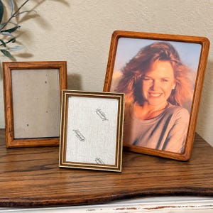 May include: Three rectangular picture frames with wooden borders. One frame on the left is empty, a smaller central frame contains a white insert with the text "Hartcraft" printed repeatedly, and a larger frame on the right holds a portrait photo.