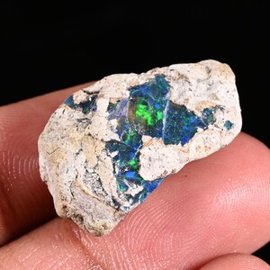 May include: A raw, unpolished opal specimen featuring a rough, light-colored, porous rock matrix surrounding a central, vibrant patch of iridescent blue and green play-of-color that catches the light with a distinct, sparkling, crystalline texture.
