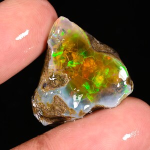 Puede incluir: Una gema de ópalo en bruto con una vibrante exhibición de color. La piedra presenta una mezcla de tonos verdes, naranjas y amarillos, con una apariencia agrietada y texturizada. El ópalo se sostiene entre dos dedos, mostrando su forma natural y en bruto.