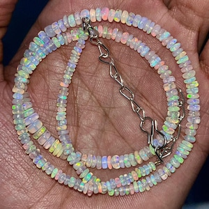 Op de afbeelding: Een delicate ketting met kleine, iriserende opaal kralen. De kralen zijn geregen met een zilveren ketting en sluiting. De opalen vertonen een reeks kleuren, waaronder wit, roze en groen, waardoor een glinsterend effect ontstaat. De ketting is ontworpen om om de nek te worden gedragen.