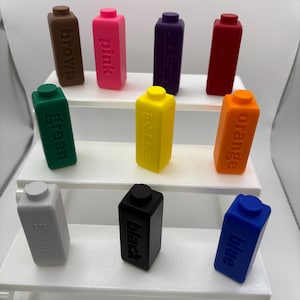 May include: A collection of nine rectangular blocks in various colours including brown, pink, purple, red, green, yellow, orange, grey, and blue. Each block features a raised circular stud on top and embossed text or braille indicating its colour.