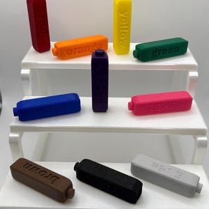 May include: A collection of ten rectangular plastic blocks in various colours including red, orange, yellow, green, blue, purple, pink, brown, black, and grey. Each block features embossed text or braille patterns on its surface.