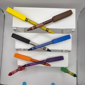 May include: A collection of Crayola crayons, each fitted with a rectangular plastic holder. The crayons are in various colors including yellow, brown, black, blue, orange, purple, red, and green, arranged in a layered display on white tiers.