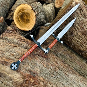 May include: A long sword and a shorter dagger with silver-toned steel blades, black metal crossguards, and brown wood-grain patterned handles. Both hilts feature a black metal pommel with a white cross emblem centred on the circular base.