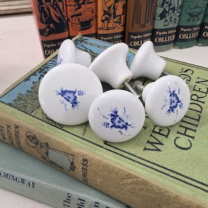 May include: A set of five white ceramic cabinet knobs, each featuring a blue floral pattern on the front. The knobs are round with a smooth finish and include metal hardware screws for installation, displayed in a group on top of books.