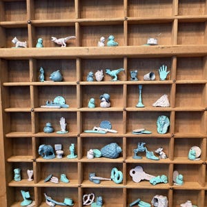 May include: A wooden printer's tray with many small compartments, each containing a variety of miniature figurines and objects in shades of light blue, teal, and white, including animals, household items, and geometric shapes arranged in rows.