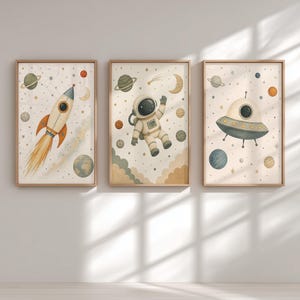 May include: Three framed wall art prints featuring space themes. The left print shows a rocket ship, the center print depicts an astronaut floating in space, and the right print displays a flying saucer, all set against a background of stars and planets.