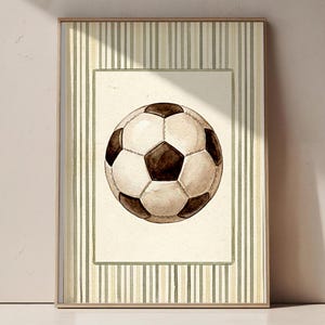 May include: Watercolor painting of a soccer ball in a frame. The ball is white with black pentagons, set against a cream background. The frame has a striped pattern in shades of green and beige, with a light brown border.