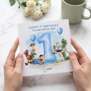 May include: A square birthday greeting card featuring a large blue watercolour number one, surrounded by Toy Story characters including Woody, Buzz Lightyear, Rex, Bullseye, Mr. Potato Head, and an Alien. Text reads: HAPPY 1st BIRTHDAY TO OUR SPECIAL SON.
