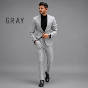 May include: A light gray suit consisting of a single-breasted blazer with notched lapels and matching slim-fit trousers. The suit is worn over a black mock-neck shirt. The word GRAY is printed in dark letters on the left side of the image.