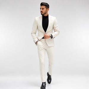May include: A cream-colored suit consisting of a single-breasted blazer with notched lapels and matching slim-fit trousers. The suit is worn over a black mock-neck shirt, paired with black leather dress shoes and a silver-toned wristwatch.