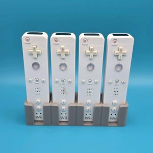 May include: Four white video game controllers with a rectangular shape, each featuring a power button, a directional pad, an A button, a home button, plus and minus buttons, and numbered 1 and 2 buttons, all standing in a gray plastic charging dock.