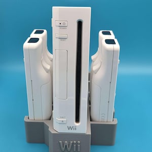 May include: A white Wii game console positioned vertically in a gray 3D-printed stand, flanked by four white Wii remote controllers. The console features power, reset, and eject buttons, with the Wii logo printed on the front and the base.