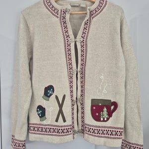 May include: A beige knit cardigan featuring a round neckline and long sleeves with maroon and dark green patterned trim. The front displays embroidered dark green mittens, brown skis, and a maroon mug with white steam and small tree designs.