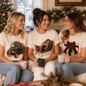 Three smiling women sit together on a couch, each wearing a beige t-shirt printed with a different vintage Christmas illustration and holding a mug.