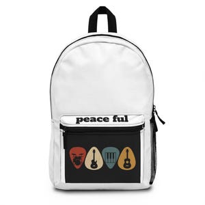 Puede incluir: Mochila blanca con un bolsillo frontal negro con el texto "peace ful". El bolsillo muestra cuatro diseños de púas de guitarra de colores: una batería, una guitarra, un teclado y una guitarra. La mochila tiene un asa y cremallera negras.