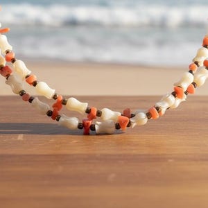 May include: A double-strand necklace featuring white carved shell beads shaped like small flowers, interspersed with irregular orange coral chips and small dark metallic spacer beads, creating a textured and layered appearance.