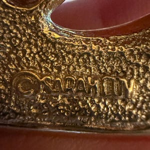 May include: Close-up of a gold-toned brooch with a textured surface. The brooch has an open design with the text "SARAH COV" embossed on the surface. The brooch is likely vintage.
