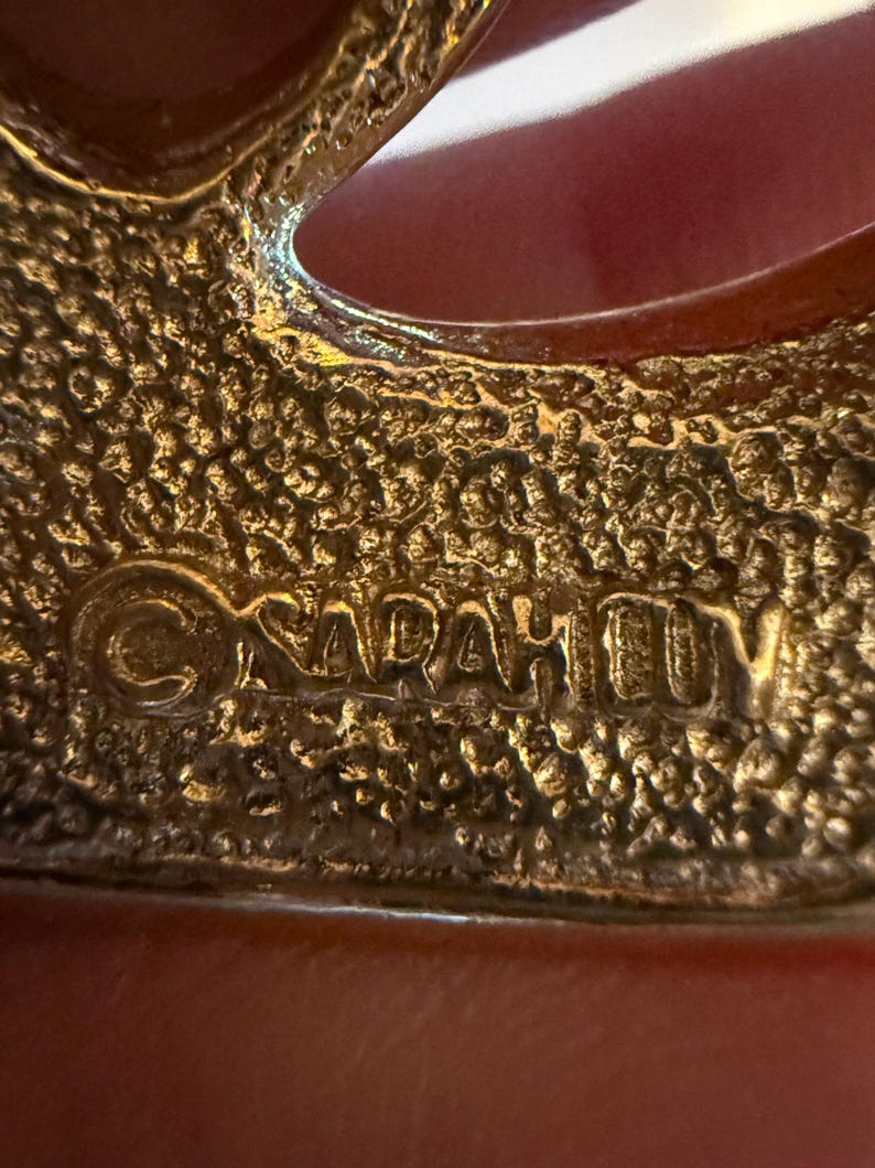 May include: Close-up of a gold-toned brooch with a textured surface. The brooch has an open design with the text "SARAH COV" embossed on the surface. The brooch is likely vintage.