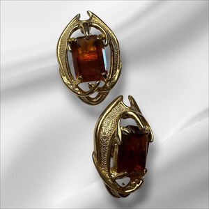 May include: A pair of vintage gold-tone clip-on earrings. Each earring features a rectangular, amber-colored stone set within an ornate, swirling gold frame. The earrings have a unique, stylized design.