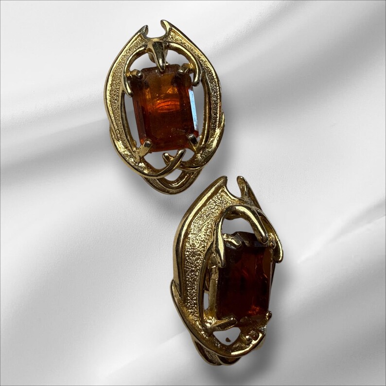 May include: A pair of vintage gold-tone clip-on earrings. Each earring features a rectangular, amber-colored stone set within an ornate, swirling gold frame. The earrings have a unique, stylized design.
