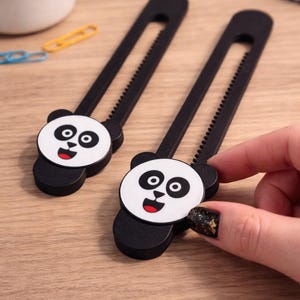 Könnte beinhalten: Zwei schwarz-weiße Panda-Schranksicherungen. Jedes Schloss hat ein Panda-Gesicht am Ende und eine gezahnte Kante. Die Schlösser sollen verhindern, dass Kinder Schränke und Schubladen öffnen. Eine Hand hält eines der Schlösser.