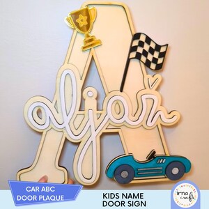 Può includere: Targa in legno per porta con il nome "Ajar" in corsivo, su una grande lettera "A". La targa è decorata con un trofeo dorato, una bandiera a scacchi e un'auto da corsa blu. Il testo sulla targa recita "CAR ABC DOOR PLAQUE" e "KIDS NAME DOOR SIGN".