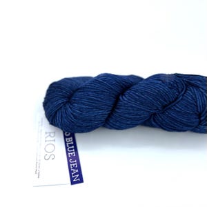 May include: A skein of dark blue yarn labeled "BLUE JEAN" and "RIOS". The yarn is tightly twisted and appears soft. The label is white with blue text and a blue border. The yarn is likely made of merino wool.