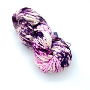 May include: A skein of thick, hand-dyed yarn featuring a variegated color pattern of deep purple, magenta, and cream. The yarn has a soft, twisted texture with distinct color transitions throughout the fiber, creating a marbled appearance.