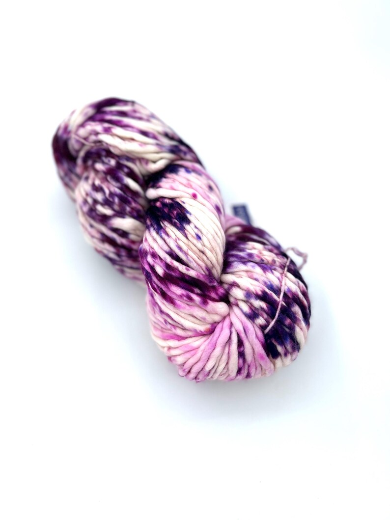 May include: A skein of thick, hand-dyed yarn featuring a variegated color pattern of deep purple, magenta, and cream. The yarn has a soft, twisted texture with distinct color transitions throughout the fiber, creating a marbled appearance.