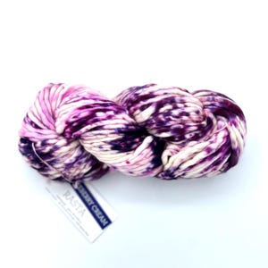 May include: A skein of thick, hand-dyed yarn featuring a variegated pattern of deep purple, magenta, and cream white. A white paper label is attached to the yarn, displaying the text RASTA and BERRY CREAM in black lettering.