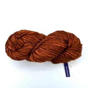 May include: A large skein of chunky, rust-colored yarn. The yarn is tightly twisted, showing a textured surface. The color is a warm, earthy tone, ideal for knitting or crocheting projects. A small tag is attached.