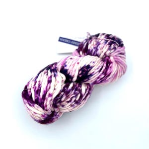 May include: A skein of thick, hand-dyed yarn featuring a variegated pattern of deep purple, magenta, and cream white. The yarn has a soft, twisted texture and includes a small white paper tag with dark blue text attached to the top section.